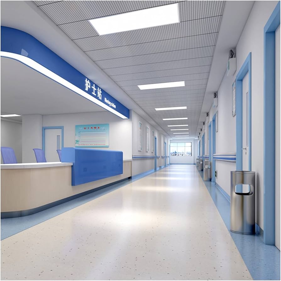 Hospital corridor with clean white and blue walls, modern ceiling lights, and a reception area in the foreground. The floor is polished, and the hallway is well-lit, leading to a distant window.