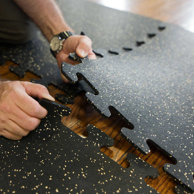 Person assembling black interlocking rubber flooring tiles with specks, placing them together on the floor.