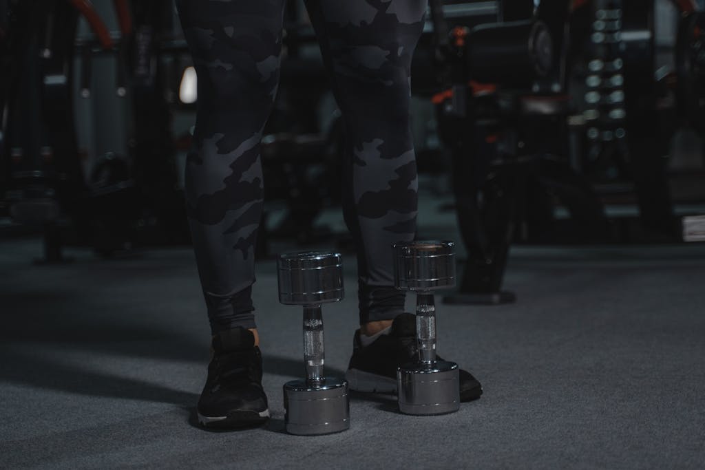 A fitness enthusiast stands with dumbbells, ready for a workout in a dimly-lit gym.