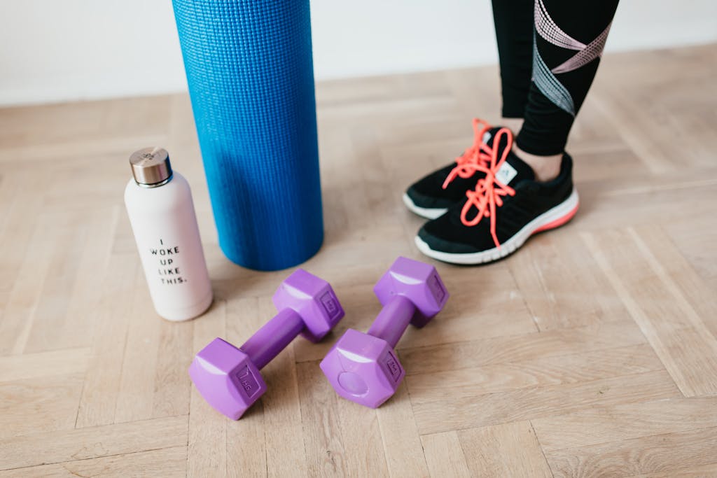 A pair of sneakers, purple dumbbells, yoga mat and bottle on wooden floor.