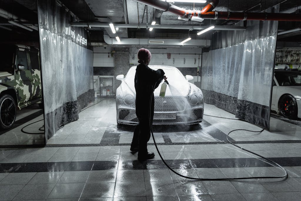 A person using a hose to spray a car with snow foam in an indoor carwash setting.