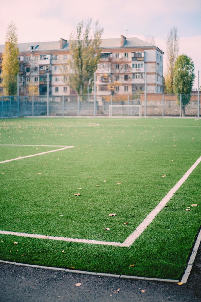 An empty soccer field with artificial turf and apartment buildings in the background, under a white sky.