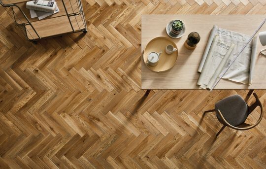 Close-up of a herringbone-patterned wooden floor with a modern table and chair.
