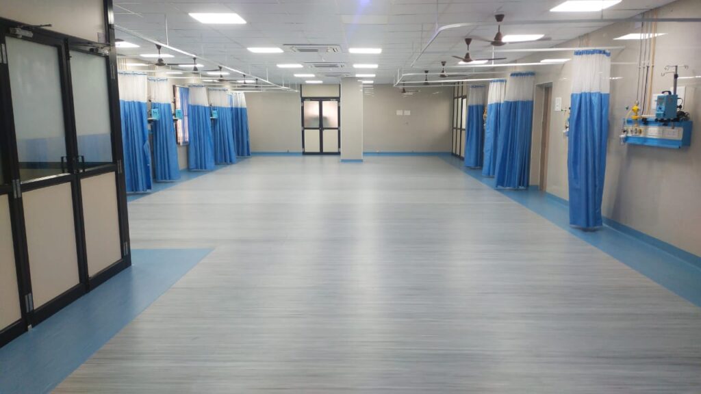Spacious hospital ward with light grey and blue vinyl flooring, privacy curtains, and modern medical facilities.